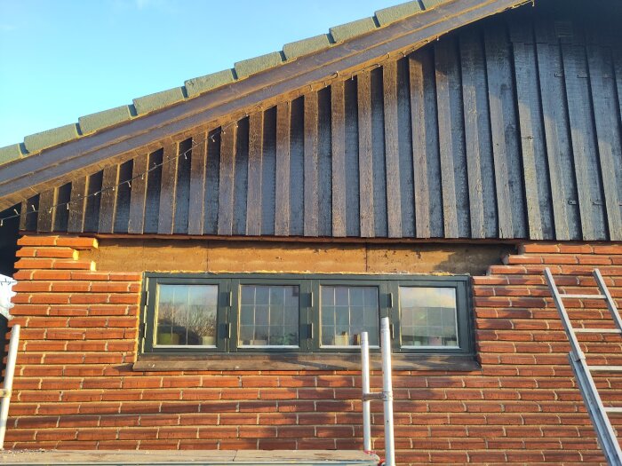 Tegelvägg med ett borttaget fönster, delar av väggen borta, en stege står lutad mot väggen, dagsljus belyser byggområdet.