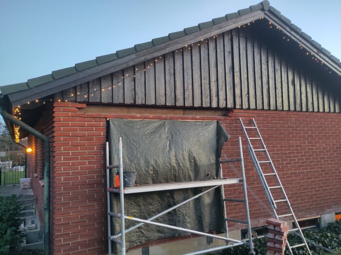 Tegelvägg med byggställning och täckt fönsteröppning, pågående renovering. Liggande stege bredvid, julljus längs takfoten.