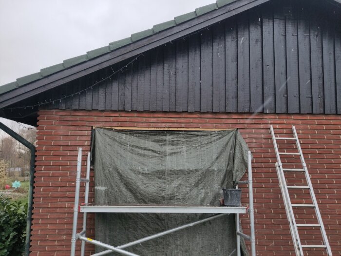 Tegelvägg med öppning för stort fönster, skyddad av presenning. Stegar och byggställning kan ses framför väggen.