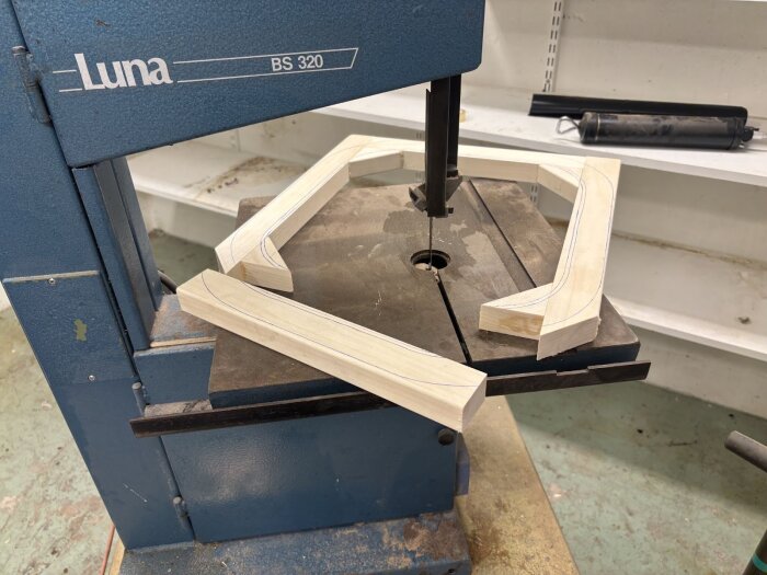 Aspträbitar för fönsterramar ligger bredvid en bandsåg i en snickeriverkstad, redo för montering och bearbetning med exakta konturer.