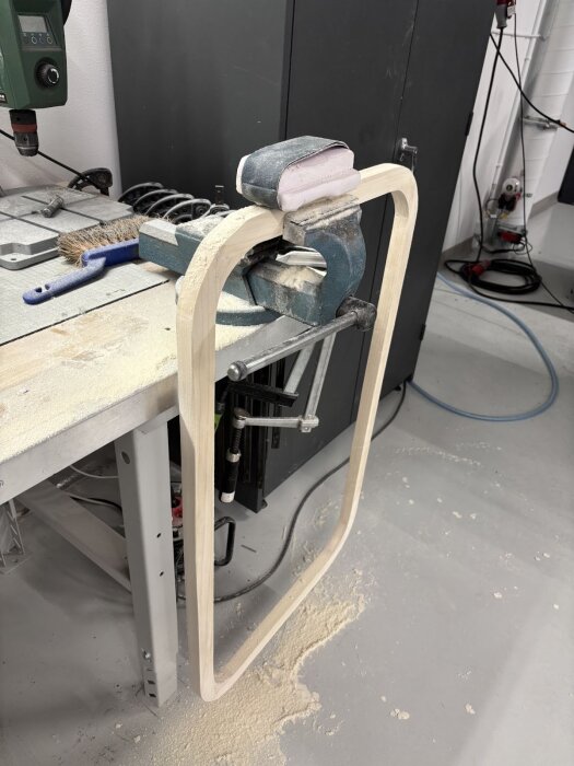 En träram formas i en verkstad, placerad i ett skruvstäd på en arbetsbänk. Slipdamm syns runt arbetsstationen.