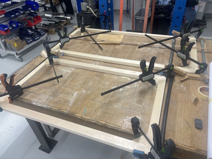 Träramar för fönster spärras under limmning på ett arbetsbord, omgivna av verktyg och utrustning i en verkstadsmiljö.