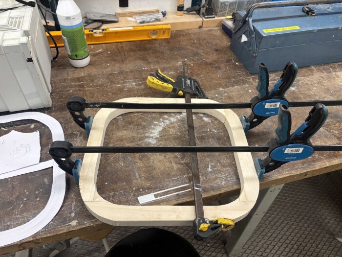 Träram fäst med tvingar under tillverkning på arbetsbänk, med mönsterpapper och verktyg i bakgrunden.