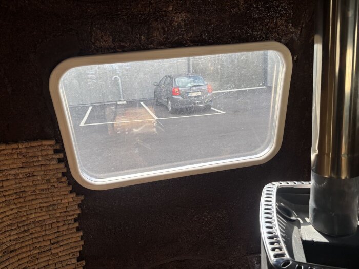 Rektangulärt fönster med vit träram invändigt, utsikt mot parkeringsplats och bil. Inredning med korkdetaljer synliga på vänster sida.