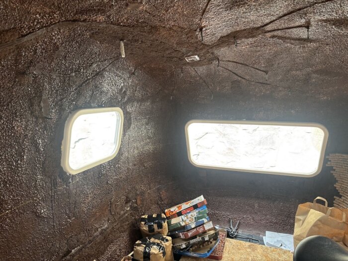 Inuti en byggnad med två rektangulära fönsterkarmar, synliga verktyg och olika lådor. Väggarna är täckta av brun kork.