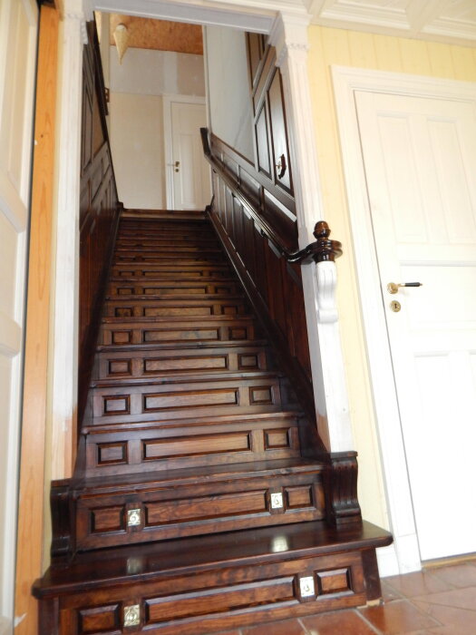 Trätrappa med mörka paneler, svarvade knoppar och pelare, samt små detaljer på stegen, på väg upp till övervåningen.