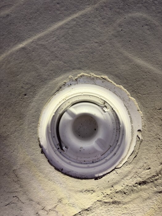 Golvyta med ett runt avloppsbrunnslock omgivet av flytspackel, där lockets omgivning är ojämn och spacklet sprucket nära brunnen.