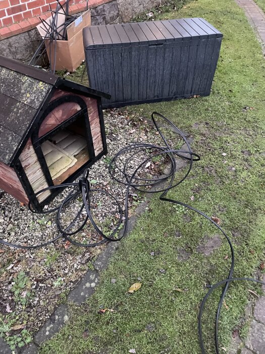 Utomhusbild med ett hundhus på grus, en plastlåda och svarta kablar på gräsmattan vid en tegelvägg.