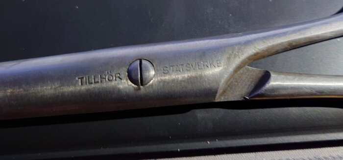Närbild på handtaget av en rostig sax med texten "TILLHÖR STATEN" ingraverad.