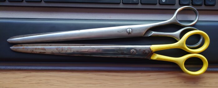 Två metallsaxar ligger på ett skrivbord, en med gula handtag och den andra i metallfärg, ovanpå ett tangentbord och musmatta.