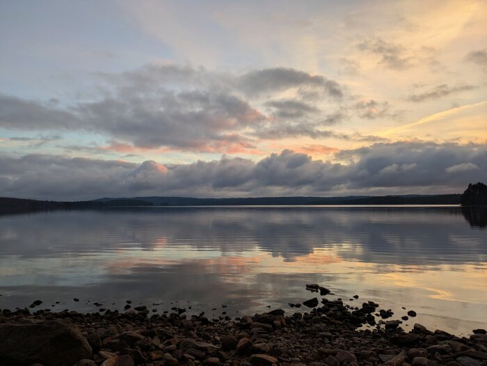 Solnedgång över en stilla sjö där himlen speglas i vattnet, omgiven av stenig strand och barrskog i horisonten.