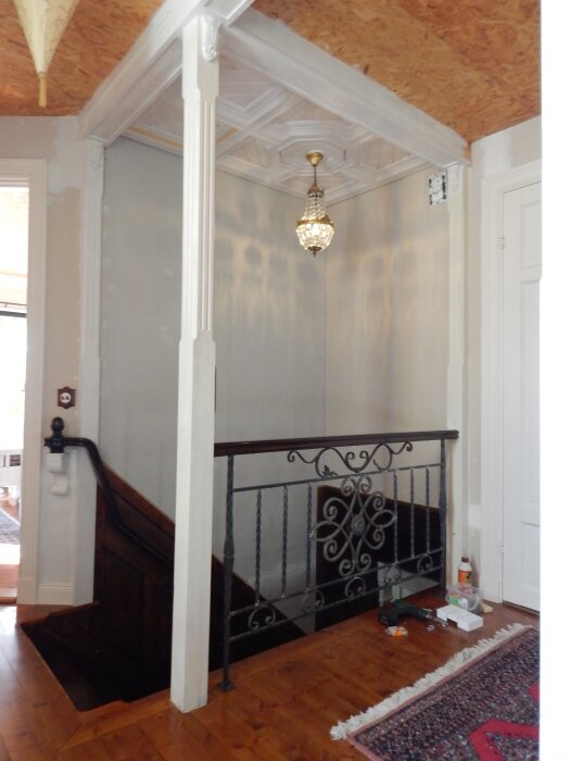 Övervåning under konstruktion med trappräcke av smide, trätrappa och lampa i taket; verktyg och material på golvet.