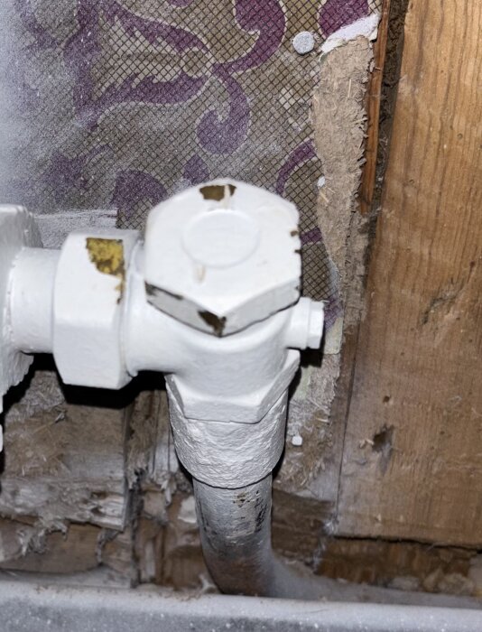 Vit radiatorventil med anslutningar på en gammal ledning, framför mönstrad tapet samt träpanel i bakgrunden.