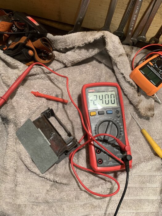 Multimeter visar 2,4 volt på en grå yta, omgiven av verktyg på en arbetsduk.