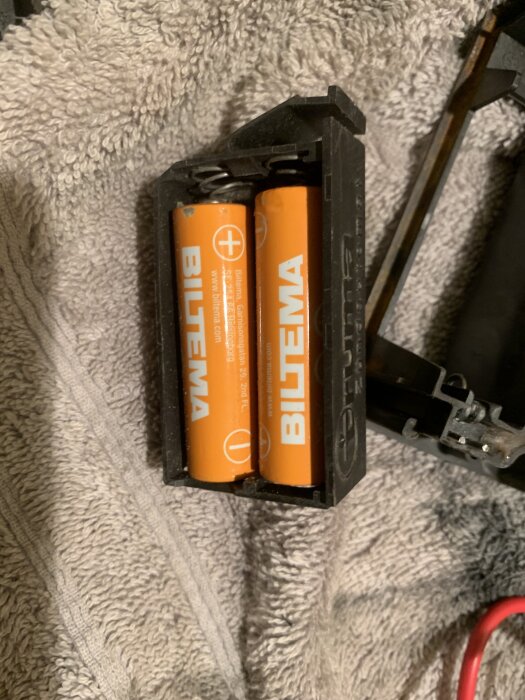 Bilden visar två orangea Biltema-batterier i en batterihållare, placerade på en grå handduk.