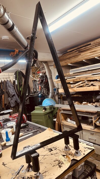 Triangulär träram med svarta sängben, placerad i en verkstadsmiljö med verktyg och material i bakgrunden.