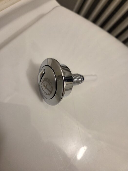 Närbild av en Gustavsberg spolknapp med silverfärgad finish, placerad på en vit yta.