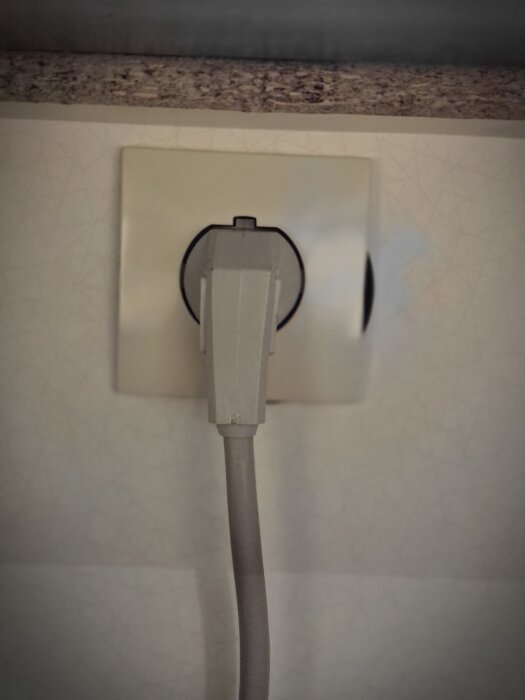 Grå kontakt som sitter i ett vägguttag under en köksbänk, kopplad till en keramikhäll, fokus på strömanslutningen.