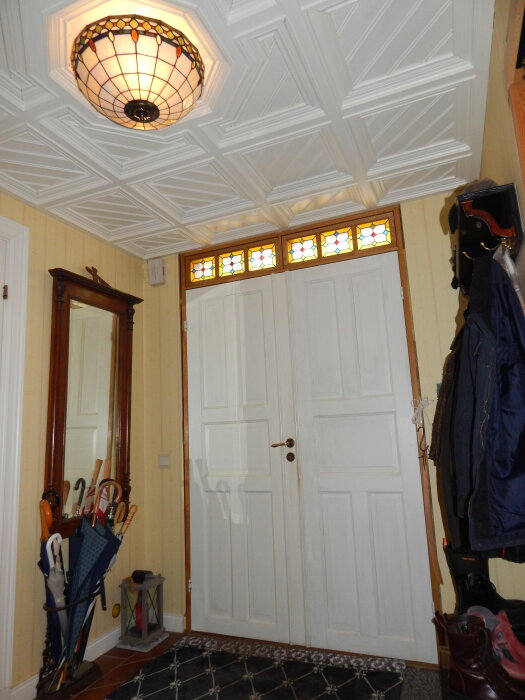 Vit hall med egentillverkade fönster, vitmålat paneltak, spegel, paraplyställ, och stövlar vid en vit ytterdörr med ofärdiga dörrfoder.