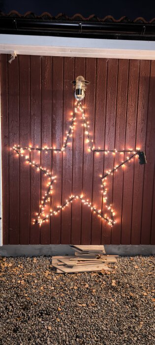 Ljusslinga formad som en stjärna monterad på en röd fasadvägg, med en utomhuslampa ovanför och en hög med plankor på marken nedanför.