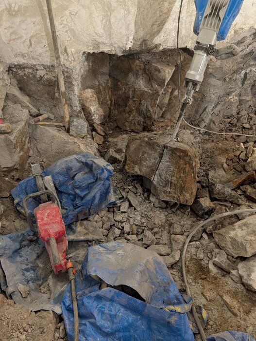 Byggarbetsplats i källare med stenhögar. En brytmaskin borrar i klippväggen omgiven av verktyg och blå presenningar på marken.