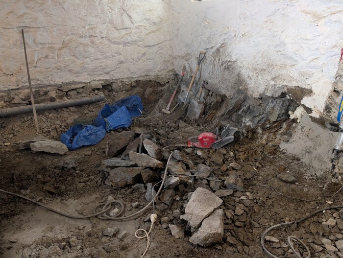 Utgrävning i källare med stenskräp, blå presenning, verktyg och en såg; synliga väggar och ledning.