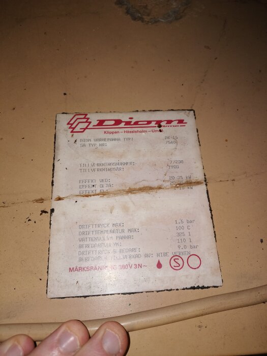 Etikett på Diom värmepanna, visar tekniska specifikationer som typ, serienummer och driftdata. Bakgrunden är brun och en hand syns i nederkanten.