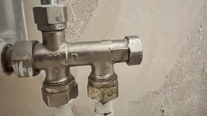 Metallisk Danfoss-ventil monterad på ett rörsystem, märkning 87B syns, installerad med kopplingar framför en vägg med textur.