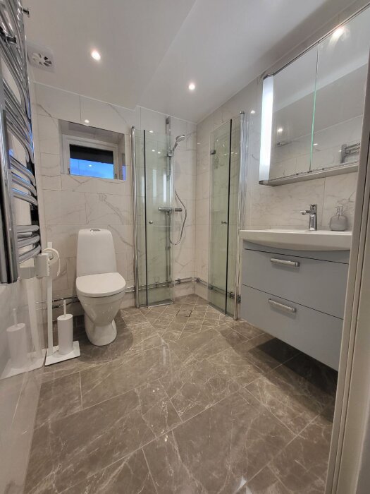 Modernt badrum med kaklade väggar och golv, duschkabin, toalett, handfat med spegelskåp samt grå skåp och handduksvärmare.