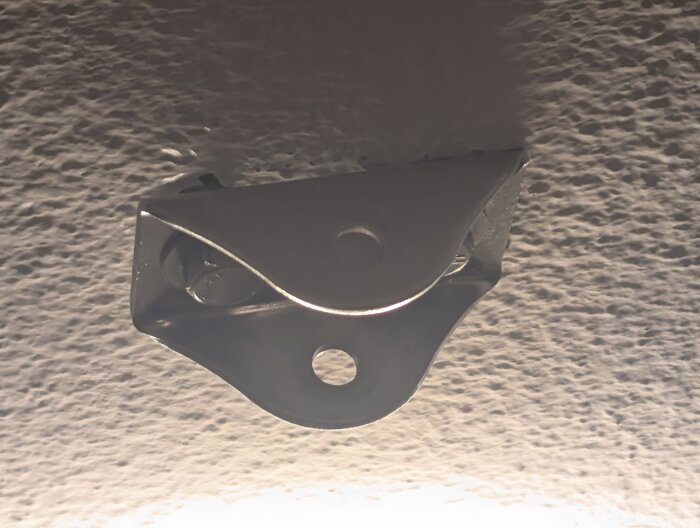 Metallfäste för gymnastikringar monterat i taket. Två bultar synliga i ett kornigt betongtak, en del av ett hyreshus från 60-talet.