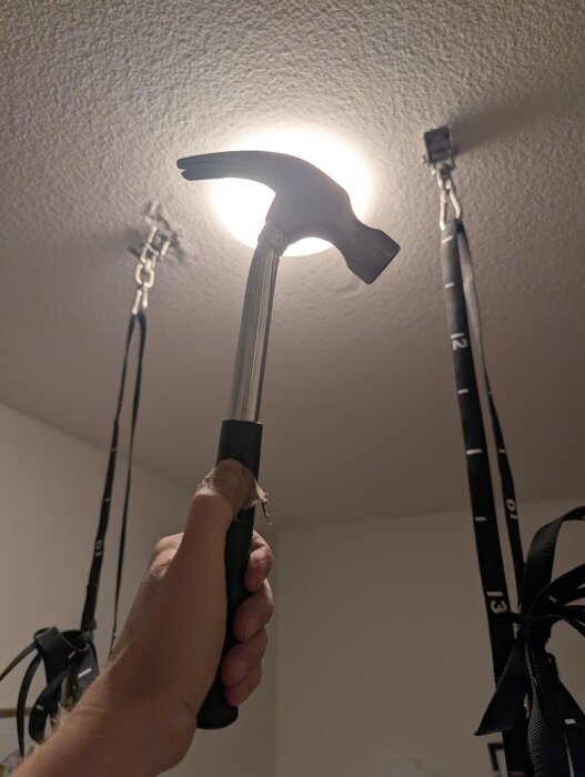 Hand håller hammare under takfästen med upphängda gymnastikringar i ett rum, ljuskälla i taket.