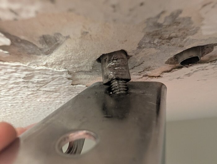Metallfäste med en bult i taket på 60-talshus, där bulten är delvis urskruvad men fastnar och omgivande färg flagar.