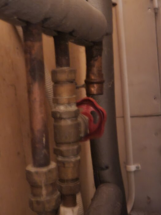 Skjutventil i kopparledningar med röd ventilratt, del av äldre vattensystem i behov av byte.