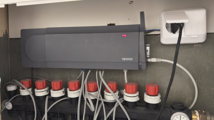 En Uponor centralenhet med anslutna kablar och rör i ett skåp, med röda vridknappar synliga nedanför.