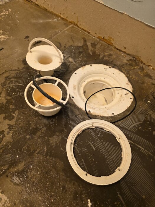 Demonterade delar av golvbrunn på betonggolv, inkluderar lock, vattenlås och brunnsringar; brunnen redo för fortsatt renovering.