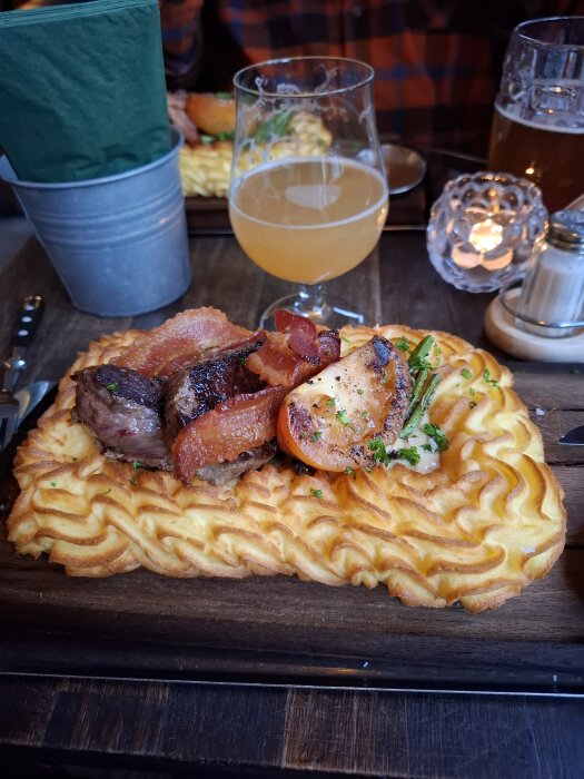 Plankstek serverad med potatismos, kött, bacon och grönsaker på en träplanka, med ett glas IPA i bakgrunden.