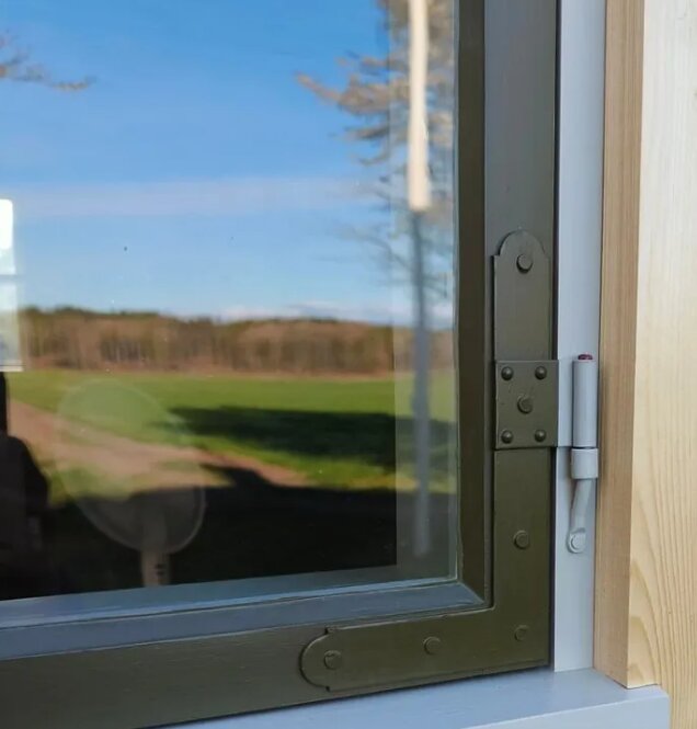 Fönster med utanpåliggande gångjärn och gångjärnsstaplare, omgärdat av trä och med en lantlig utsikt i bakgrunden.