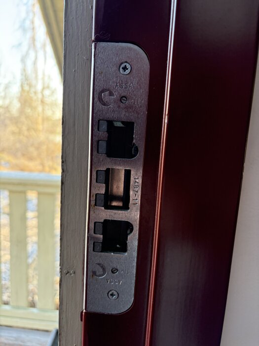 ASSA 2002 dörrlås och 1487-11 slutbleck på en dörrkarm, utomhusmiljö i bakgrunden.