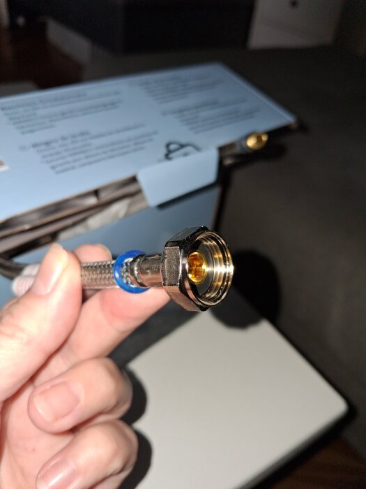 Närbild på en hand som håller en anslutningsslang med mutter för tvättställsblandare i storlek G3/8, mot en suddig bakgrund.