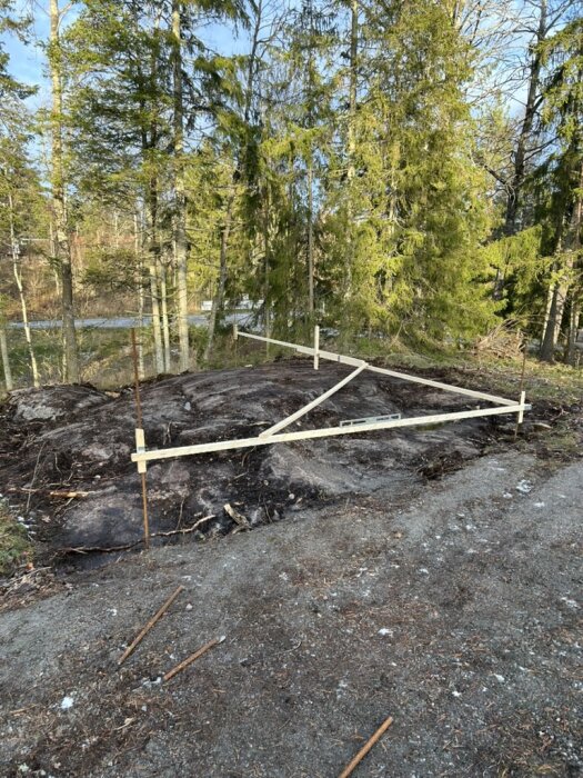 Uppmätt plats med snören och pinnar på en ojämn bergsyta omgiven av skog för att planera för Attefallshusbygge.