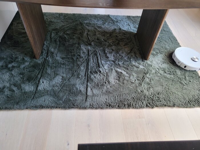 Grå matta under ett bord med en robotdammsugare på sidan. Mattan ser ojämnt rengjord ut med tydliga veck och märken.