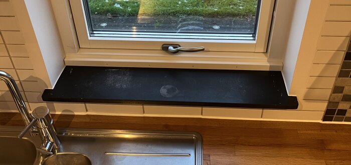 Sliten svart fönsterbräda i eternit med fläckar, framför köksfönster och diskbänk.