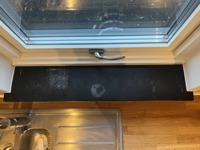 Svart, sliten fönsterbräda av eternit under ett fönster med vask och diskho i förgrunden.