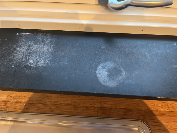 Svart fönsterbräda i eternit med fläckar och slitage, under ett fönster med vitt trä och fönsterhake i metall.