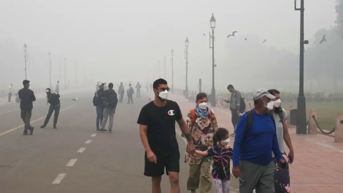 Människor promenad i dimmigt väder med ansiktsmasker i stadsområde, fåglar i bakgrunden.