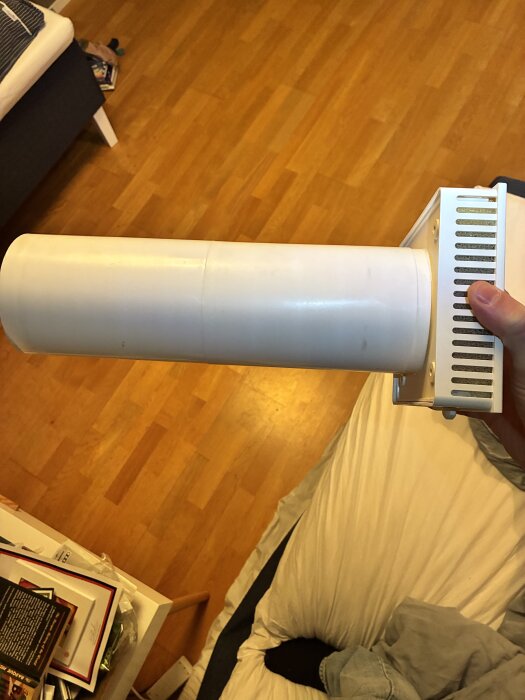 Vit plastcylinder och ventilationsgaller hålls i en hand, liggande i ett rum med trägolv och en säng i bakgrunden.