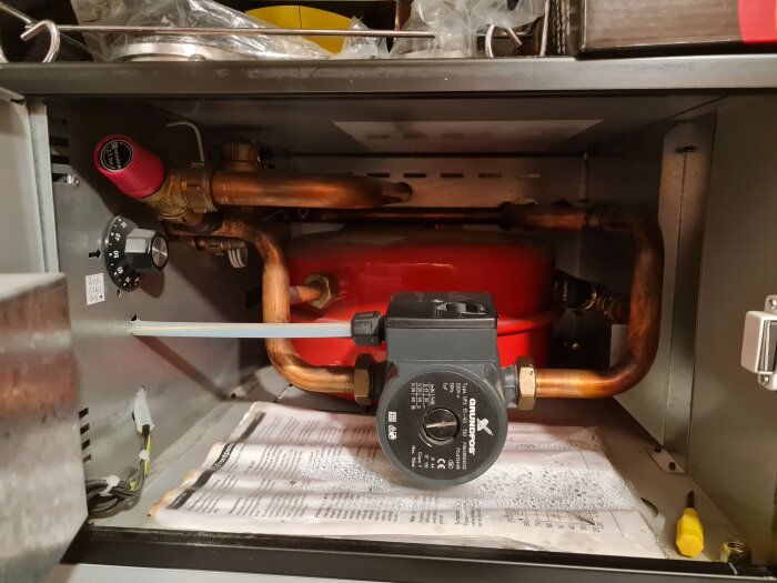 Ett vattenburet golvvärmesystem med en cirkulationspump, kopparrör och en tryckmätare syns i en öppen skåpinstallation.