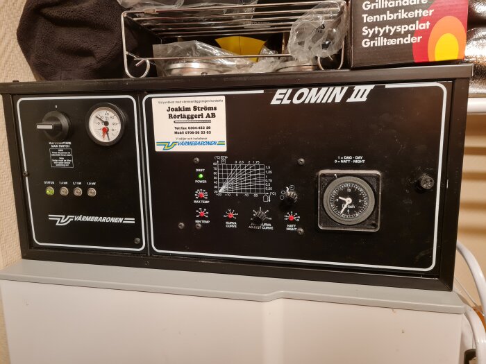 Värmepanna av märket Elomin III från Värmebaronen med tryckmätare och justeringspanel. Skylten visar kontaktinfo för rörläggare.