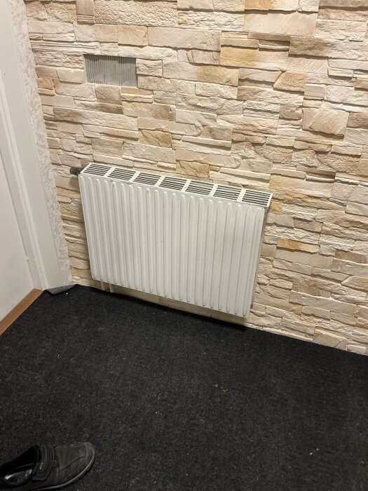 Vitt vattenburet element monterat på en vägg med beige stenimitation i ett rum med mörkt golv och en svart sko.