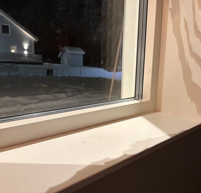 Utsikt genom ett fönster inifrån ett hus, med en isig ruta och snötäckt trädgård i bakgrunden.